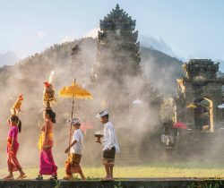 A mother and a teenaged girl are dressed in brightly colored sarongs, blouses, and sashes and are balancing tall fruit baskets on their heads. Two sons are dressed in sarongs and white shirts. The family is walking in front of an old stone temple building which has a smoky atmosphere.