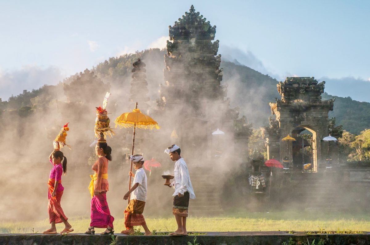 Showing traditional Balinese male and female ceremonial clothing and religious offerings as a mother and children walk to a Hindu temple (pura) in Bali.